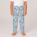 A child wearing light blue pajama pants with a repeating pattern of dogs in winter scenes, standing barefoot against a plain light gray studio background.