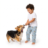 A young boy with curly dark hair wearing a white t-shirt and light blue pajama pants with a dog print, playing tug-of-war with a corgi on a white studio background.