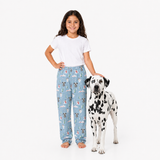 A young girl stands barefoot next to a Dalmatian dog against a plain white studio background. The girl is wearing a white T-shirt and light blue pajama pants with a repeating dog-themed print, and the dog is wearing a white T-shirt with black spots.