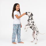 A young girl with long dark hair stands barefoot on a white studio background, smiling as she offers food to a Dalmatian dog. She is wearing a white short-sleeve T-shirt and light blue pajama pants with a dog print.
