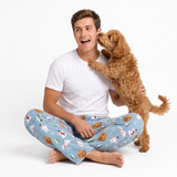 A man wearing light blue Christmas-themed pajama pants and a white t-shirt sits cross-legged on a white studio background, smiling as a fluffy brown dog rests its head on his shoulder.