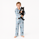 A young boy wearing a light blue pajama set with a cat-print pattern stands barefoot against a white studio background, holding a black and white cat.