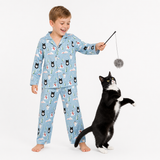 A young boy wearing light blue pajamas with a cat print stands barefoot on a white studio background, holding a wand toy while a black-and-white cat watches and reaches toward it.