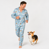 A man wearing a light blue long-sleeve pajama set with a Christmas-themed print of Santa Claus, snowmen, and snowflakes, walking barefoot next to a small corgi dog on a white studio background.
