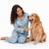 A woman in light blue pajamas with a holiday print sits on the floor next to a golden retriever dog, brushing its fur with a wooden paddle brush against a white background.