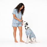 A woman and a Dalmatian dog wearing matching light blue pajama sets with a whimsical cartoon print, standing and sitting together on a seamless white studio background.