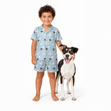 A young boy wearing a light blue short-sleeve pajama set with a cow print stands barefoot next to a black and white dog on a white studio background.