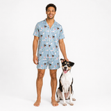 A man wearing a light blue pajama set with a dog print stands next to a black, white, and tan dog on a plain white studio background.