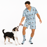 A man wearing a light blue pajama set with a dog print, including a short-sleeve button-up shirt, matching shorts, and socks, playing tug-of-war with a black and white dog on a white studio background.