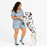 A woman wearing a light blue pajama set with a Christmas-themed print stands next to a Dalmatian dog on a white studio background. The set includes a short-sleeve button-up top and matching shorts, paired with matching socks.
