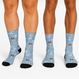 Three pairs of crew socks worn on legs, featuring a light blue background with a repeating pattern of bulldog faces, snowflakes, and small holiday motifs, with black toes and heels.