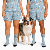 A person and a dog modeling matching light blue pajama set with a dog-print pattern, including a short-sleeve top and shorts, along with matching socks, against a plain white background.