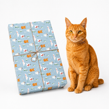 An orange tabby cat sits beside a wrapped gift box on a clean white background. The gift box is wrapped in light blue wrapping paper with a repeating pattern of white cats, orange cats, Santa Claus, and Christmas trees, tied with brown twine.