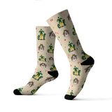 A pair of crew socks with a beige background featuring a repeating pattern of green elves, the word 'elf', and dog faces, finished with black heels, toes, and cuffs.[Socks_IconicElf]