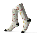 A pair of crew socks with a festive Christmas-themed print featuring Santa Claus, gifts, and the word 'Merry' on a cream background with black heels, toes, and cuffs.