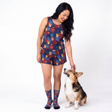 A woman wearing a matching Supergirl-themed tank top set with a  dog pattern, standing next to a corgi on a white studio background.
