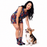 A woman wearing a Supergirl-themed printed sleeveless top and shorts set with matching socks is posing with a corgi on a white studio background.