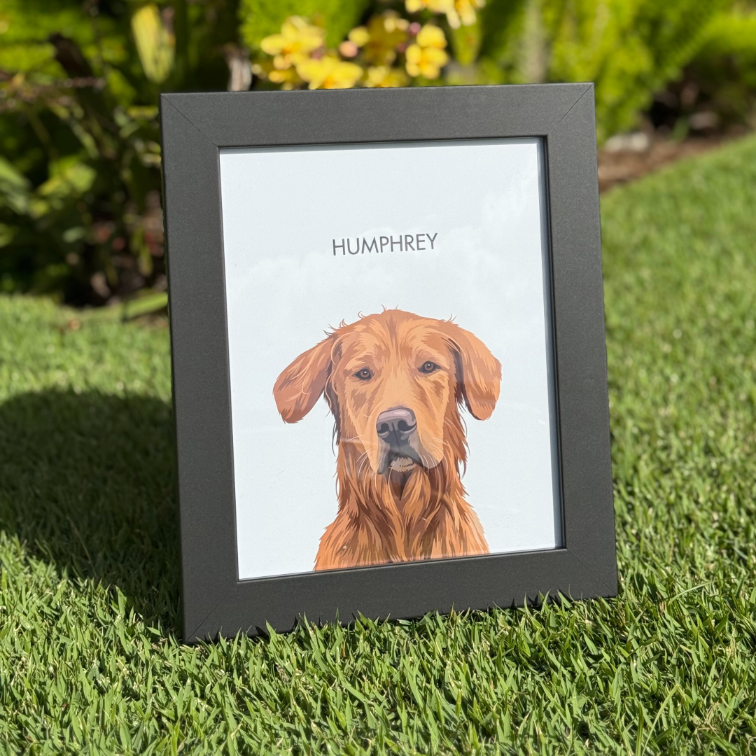 Framed illustrated portrait of a golden-colored dog labeled ‘Humphrey,’ propped upright on green grass in an outdoor garden setting with plants and sunlight in the background.