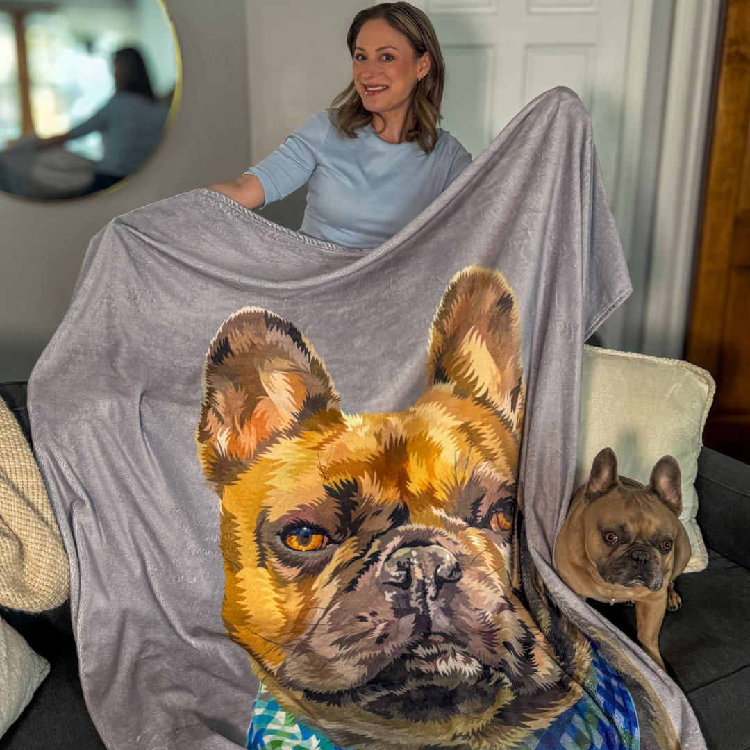 A woman sitting on a couch holds up a large gray blanket featuring a realistic illustration of a fawn-colored French Bulldog. The actual French Bulldog sits beside her on the couch, looking toward the camera.
