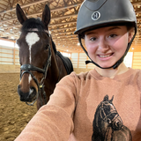 Person in helmet and horse-design sweater next to a horse in a stable.
