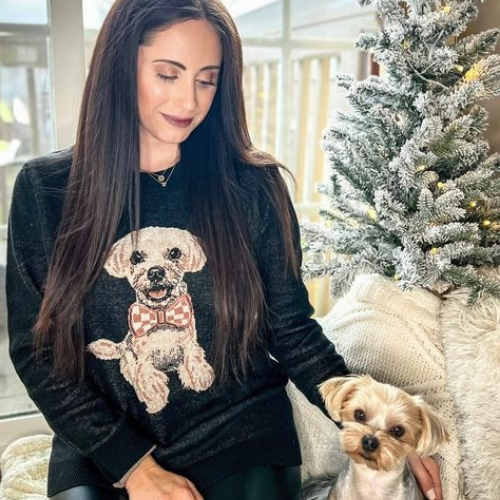 Woman wearing a sweater with a dog design, sitting next to a small dog.
