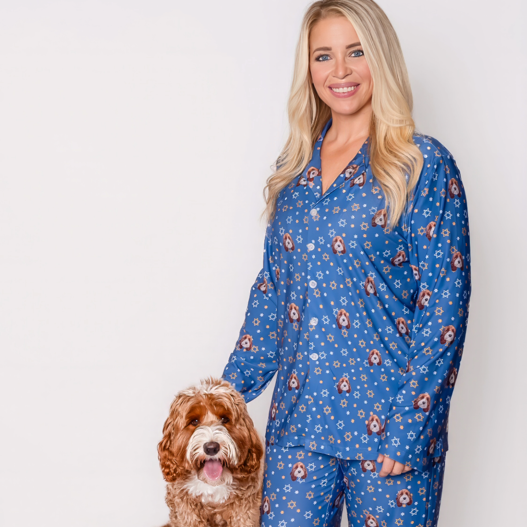 A smiling blonde woman, wearing a long-sleeve blue pajama set patterned with dog faces and white and orange Stars of David, is standing next to a medium-sized fluffy brown and white dog.