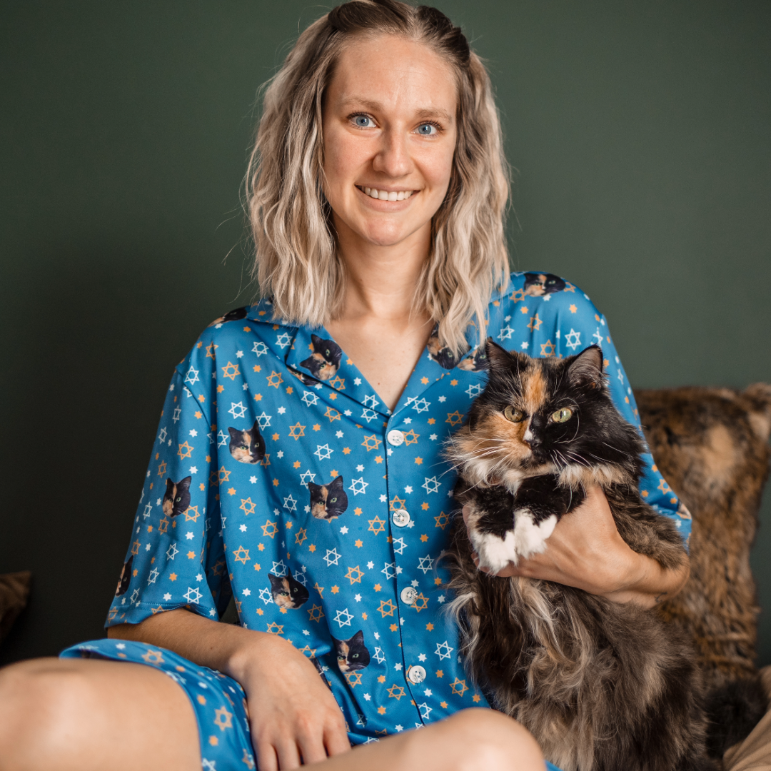 A smiling woman with blonde, wavy hair is sitting up, holding a fluffy calico cat with white paws. She is wearing a short-sleeve, button-down blue pajama set patterned with cat faces and white and orange Stars of David.