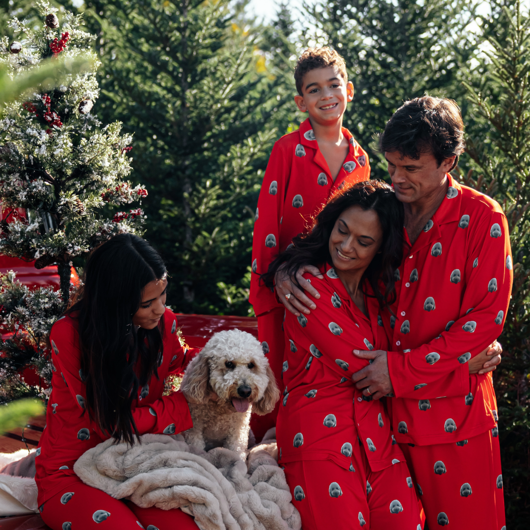 Family dressed in matching red pajamas featuring a dog face pattern, gathered outdoors among Christmas trees. A woman and man hug while a girl sits with a fluffy white dog on a blanket, and a young boy smiles standing behind them.