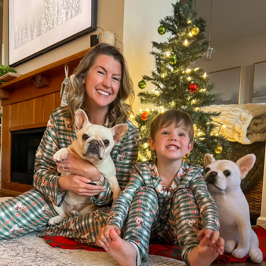 A woman and a young boy sit in front of a decorated Christmas tree, both wearing matching green plaid pajamas printed with a French bulldog pattern.