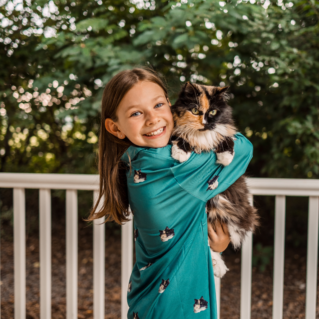 A smiling young girl wearing a teal pajama dress patterned with cat faces holds a fluffy calico cat in her arms while standing outdoors by a white railing.