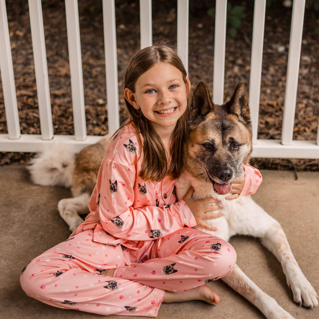 Smiling girl sitting on the ground wearing pink pajamas with dog faces, hugging a large Akita dog beside her on a porch.