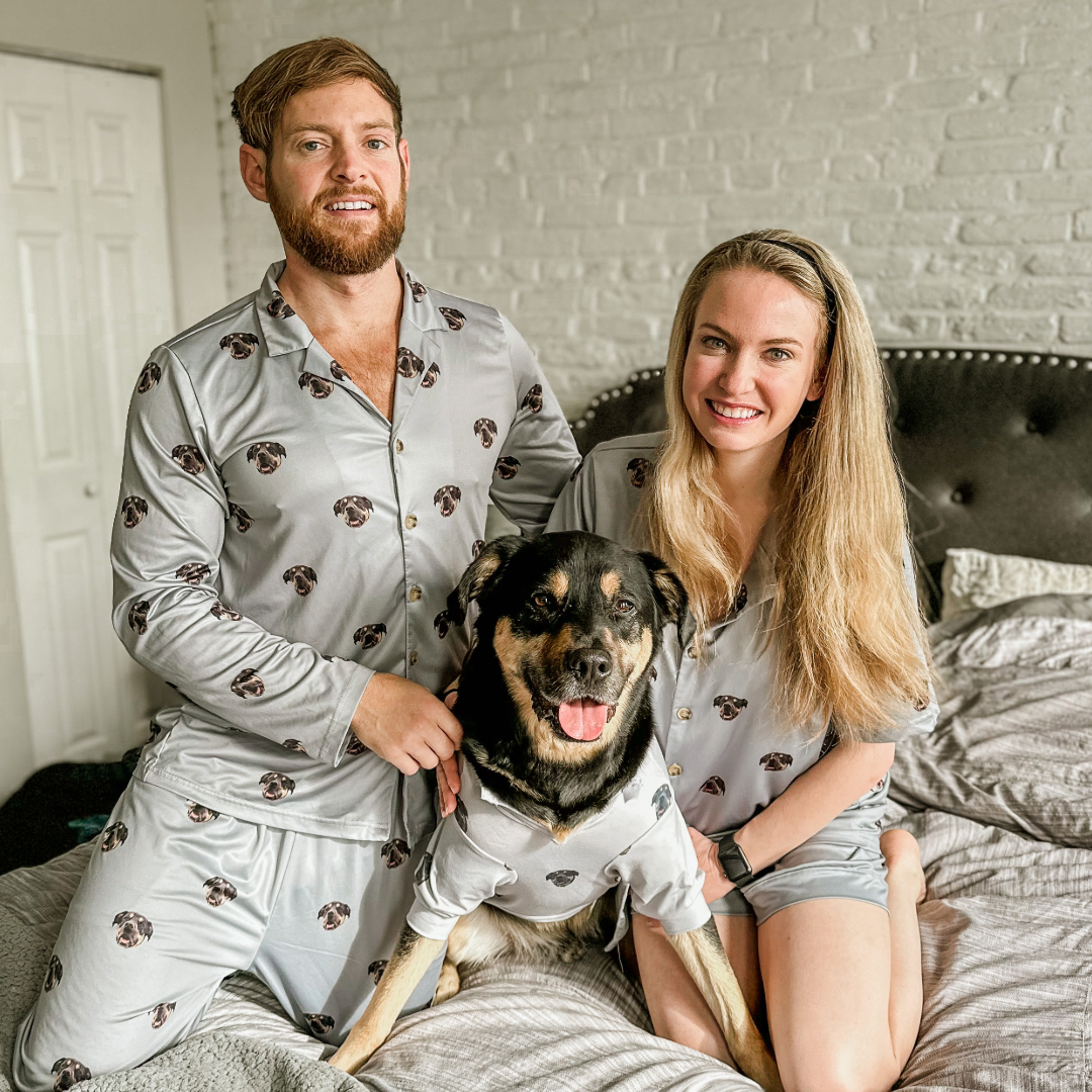 A man and woman sitting on a bed with their large black-and-tan dog between them. All three are wearing matching light gray pajamas patterned with the dog’s face.