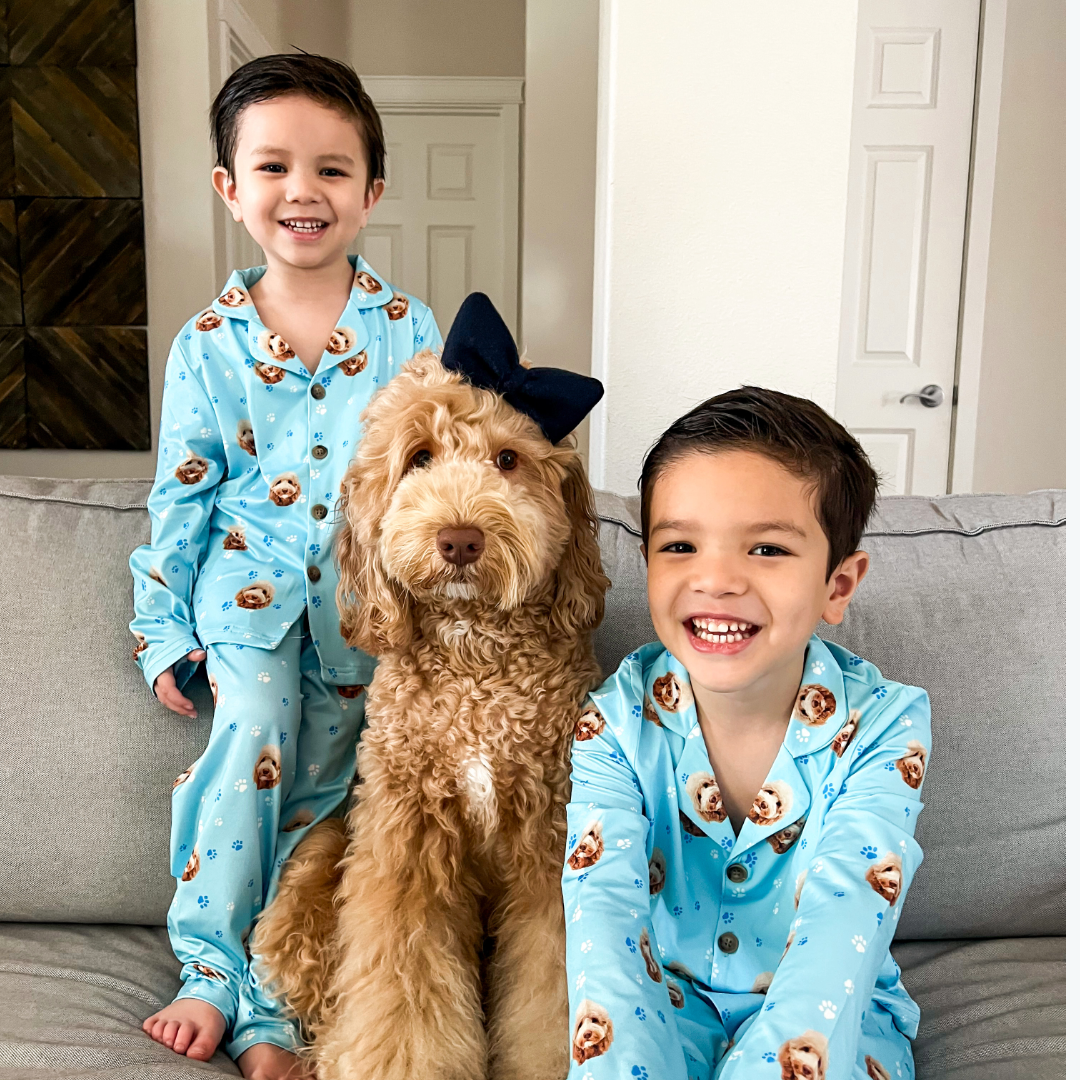 Two young boys and a fluffy light-brown dog sit together on a couch wearing matching light-blue pajamas printed with the dog’s face. One boy sits cross-legged and smiling.