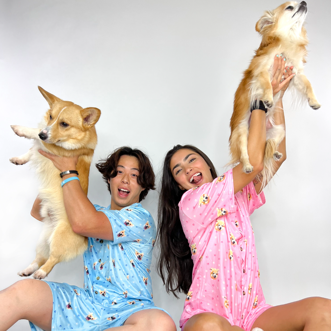 Two smiling people wearing matching pet-pattern pajama sets, each playfully lifting a small dog into the air, posed against a plain white background.