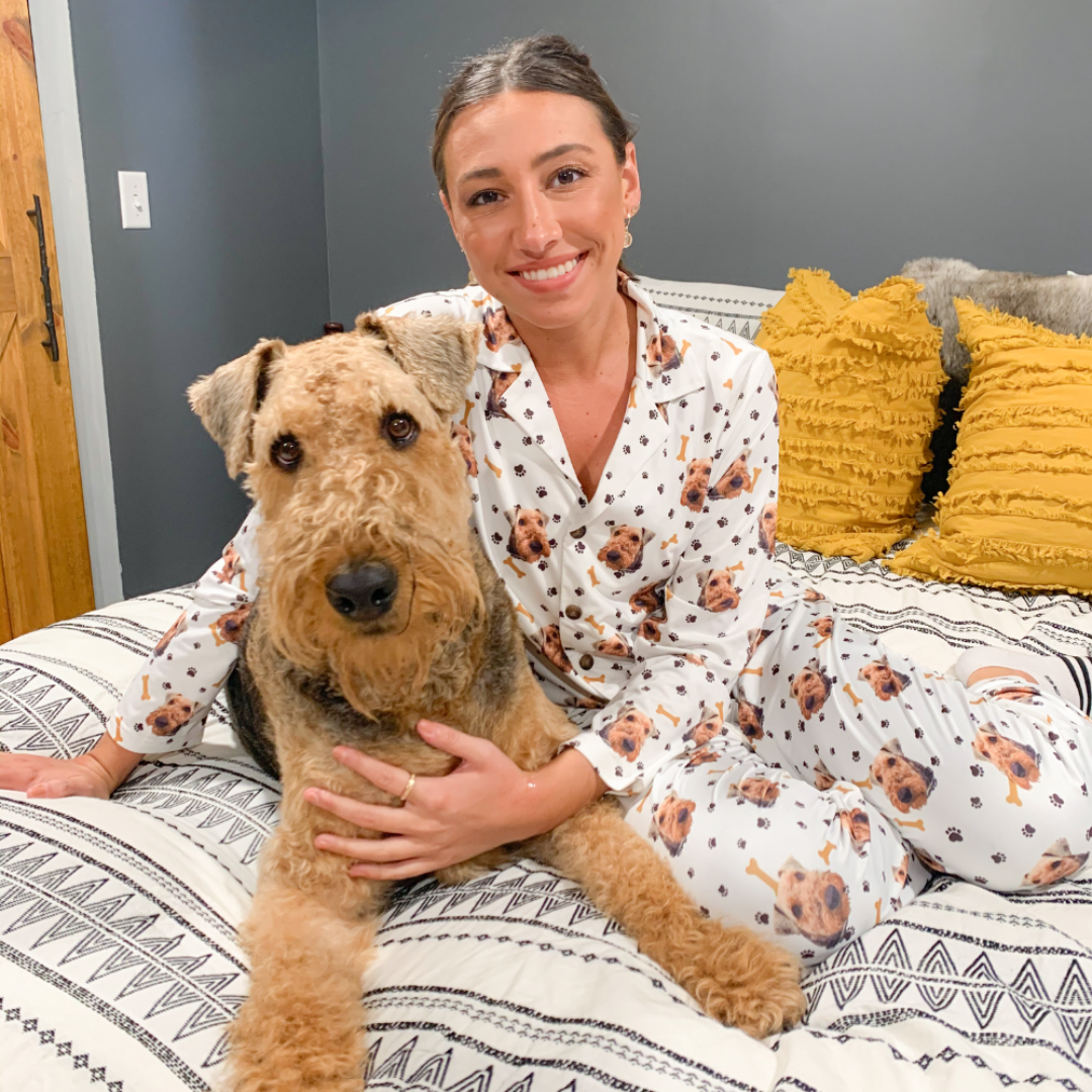 A woman sitting on a patterned bed holds a large, fluffy brown dog. She is wearing white pajamas printed with small dog faces and bones, smiling warmly at the camera.