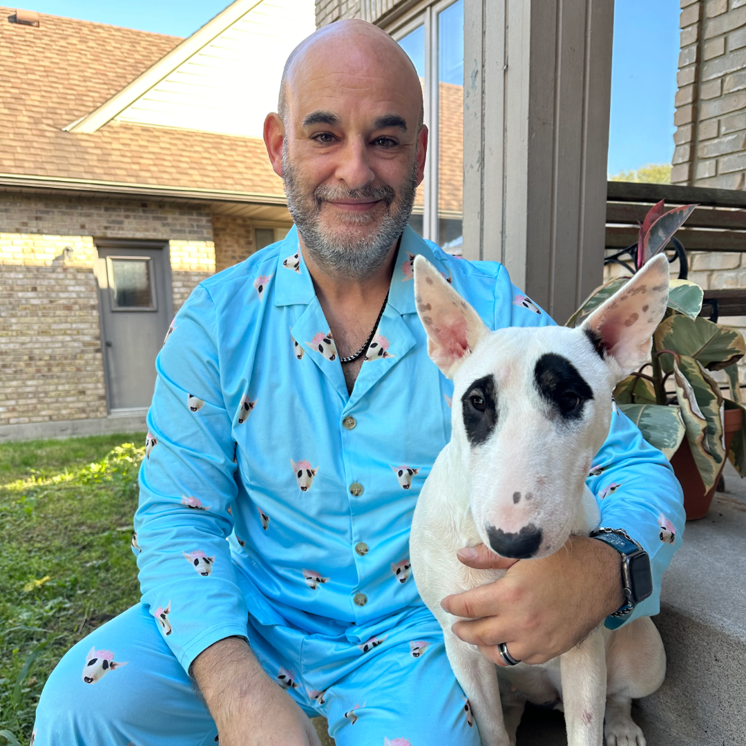 A smiling man in light blue pajamas printed with cartoon dog faces sits outside on a porch, holding a white Bull Terrier with a black patch around one eye close to him.