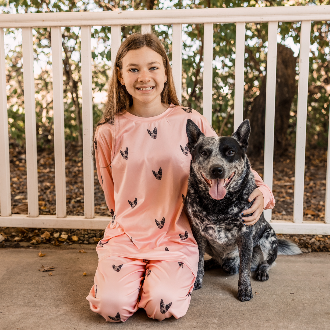 A girl wearing light pink pajamas with a repeating dog face pattern sits on a porch beside a black-and-white Australian Cattle Dog.