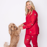 A woman wearing red pajamas printed with a dog’s face holds the front paws of a curly-haired dog who is standing upright on its hind legs. Both are facing the camera against a plain white background.