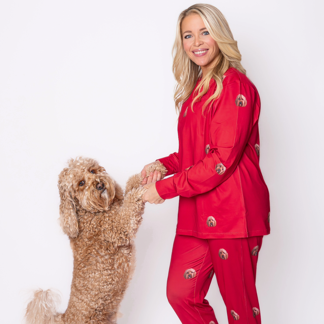 A woman wearing red pajamas printed with a dog’s face holds the front paws of a curly-haired dog who is standing upright on its hind legs. Both are facing the camera against a plain white background.
