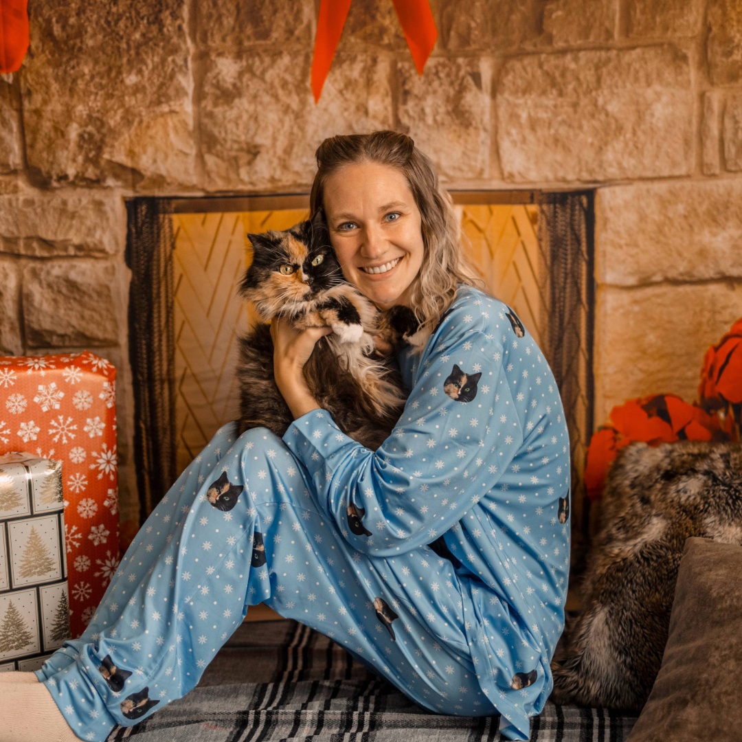 A woman sitting on a cozy holiday-themed set smiles while holding a fluffy calico cat. She is wearing light blue jogger pajamas decorated with small snowflakes and repeated cat-face prints.