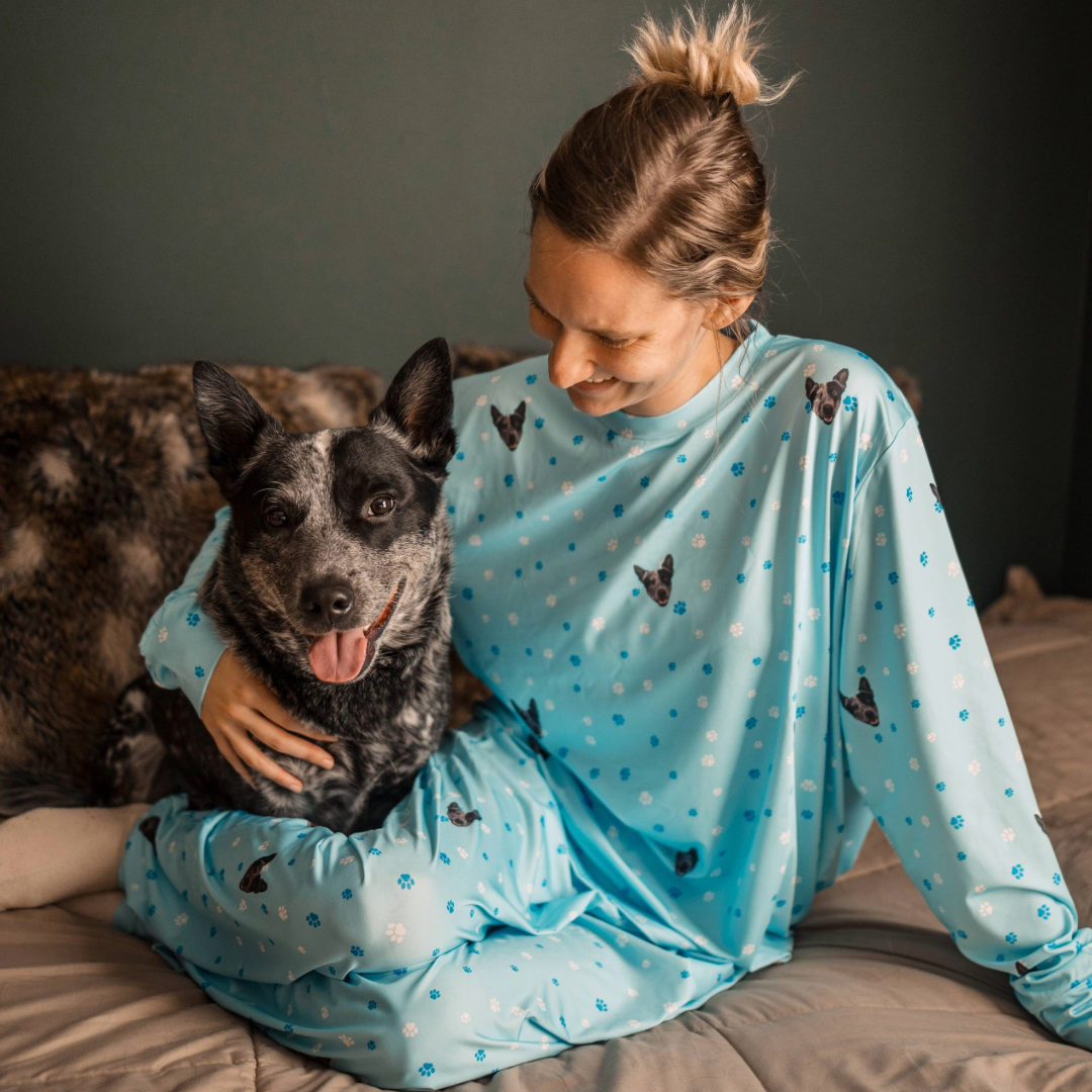 A woman sits on a bed wearing light blue jogger pajamas patterned with small dog faces and paw prints, smiling down at a happy black-and-white dog sitting beside her.