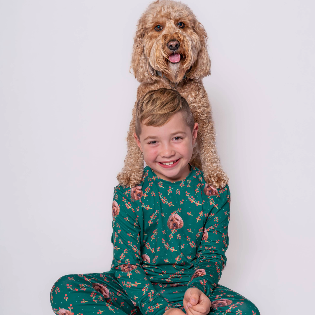 A young boy in green pajamas with a custom dog-face pattern sits cross-legged and smiling, while a curly-haired dog rests its front paws on his shoulders from behind.