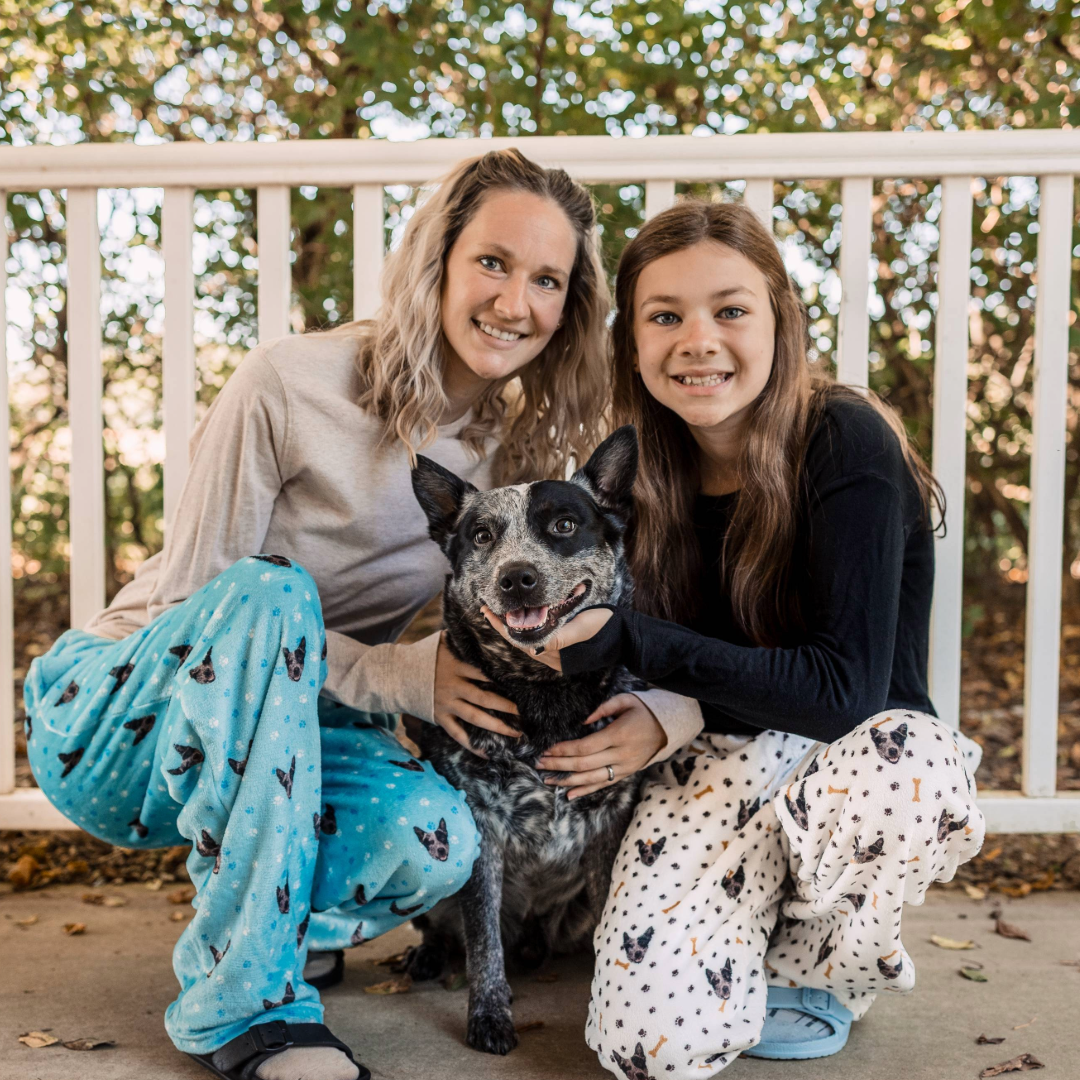 A woman and a girl kneel on a porch beside a happy black-and-white dog. Both are wearing cozy pajama pants with custom pet-face patterns, one in light blue and the other in white with small bones.