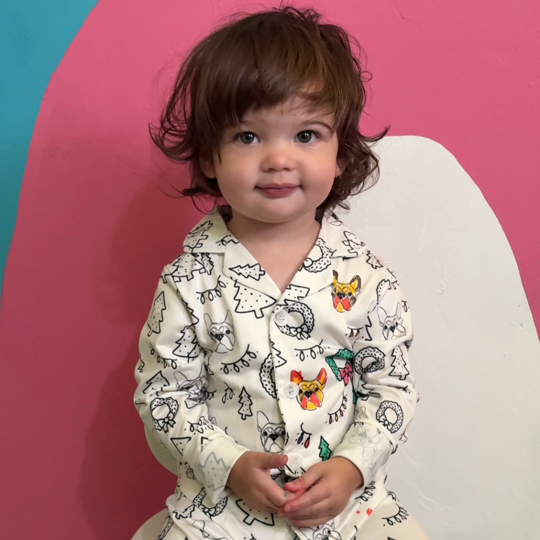 A young child with curly brown hair sits in front of a pink and teal background, wearing white colorable pajamas covered in black-outlined holiday drawings.