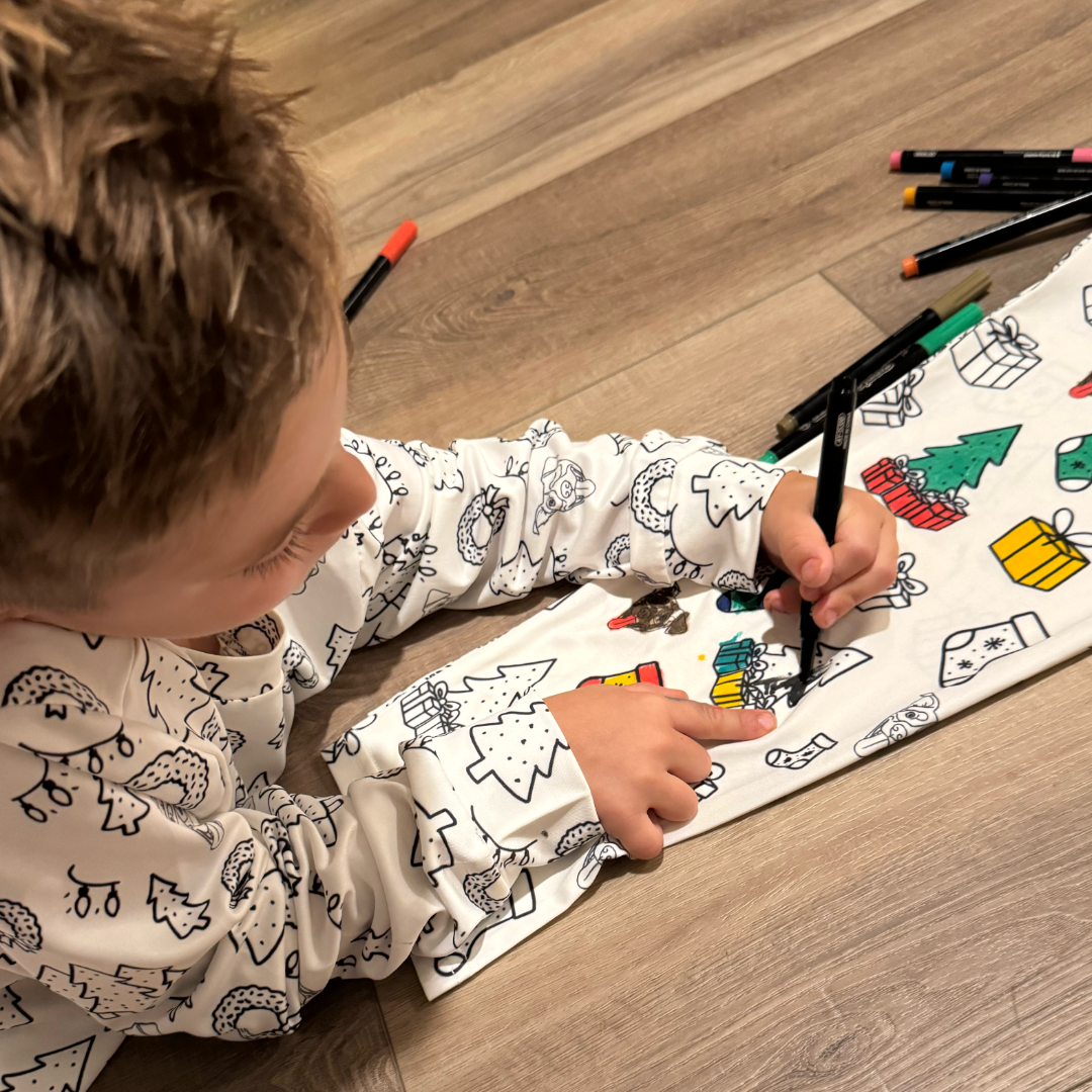 A young child lies on a wooden floor while coloring holiday-themed drawings printed on white pajamas. The pajamas feature black-outlined Christmas trees, wreaths, stockings, and dog faces.