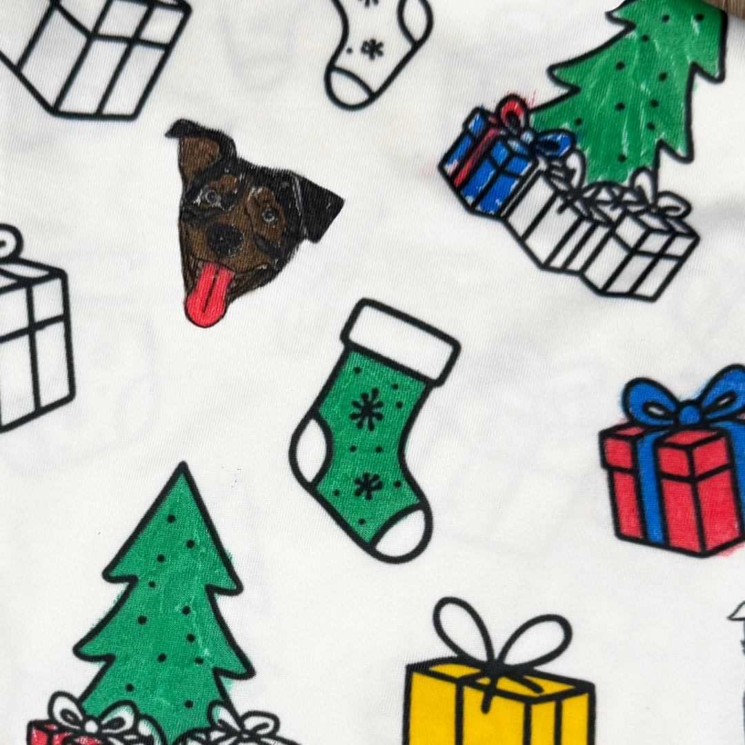 Close-up of white fabric with a playful holiday print featuring cartoon gift boxes, Christmas trees, green stockings, and a small illustrated dog face with its tongue out.