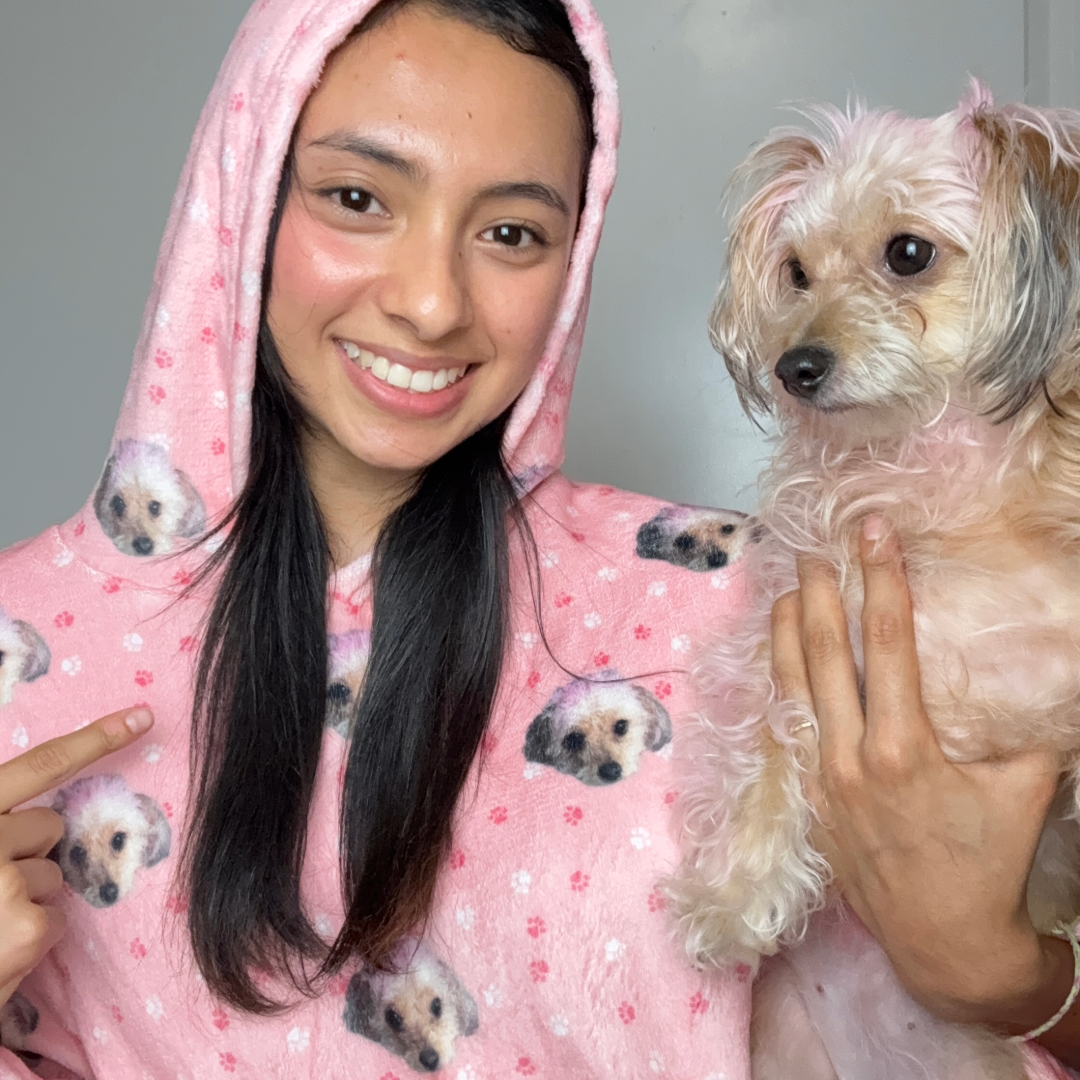 A woman wearing a pink hooded blanket covered in a repeating pattern of her small dog’s face smiles at the camera while pointing at the design. She is holding her light-colored, curly-haired dog in her other arm.