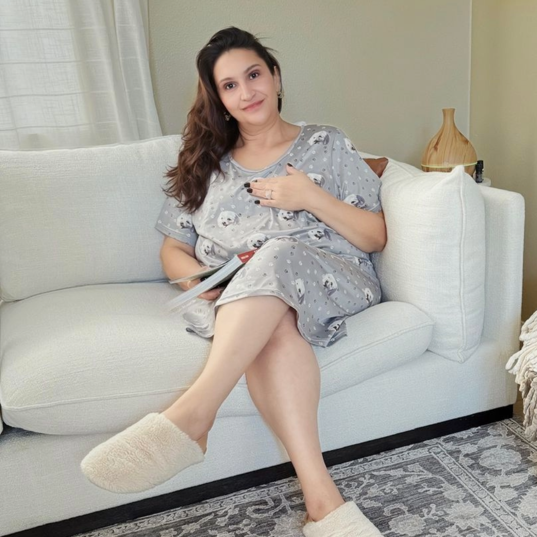 A woman sitting comfortably on a light-colored sofa, wearing a gray pajama dress printed with small dog faces and polka dots.