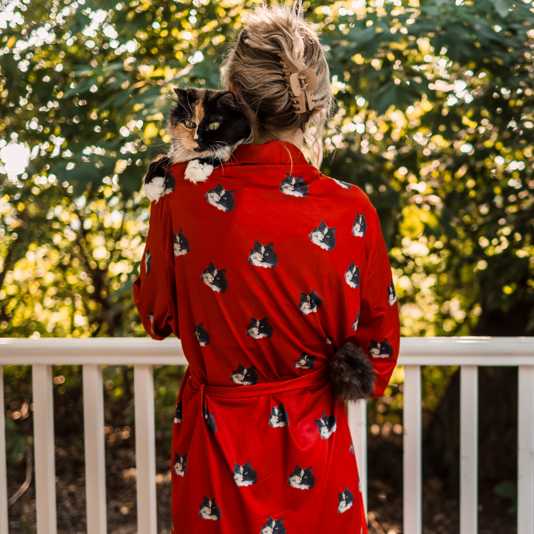 A woman standing outdoors with her back to the camera, wearing a bright red robe printed with multiple images of a black-and-orange cat’s face. She is holding a real calico cat over her shoulder.