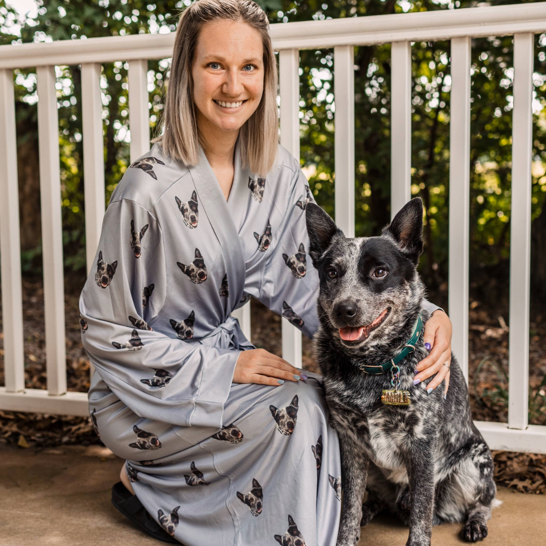 A woman kneeling on a porch while wearing a light gray robe printed with repeated portraits of her dog’s face, smiling as she poses next to her black-and-white speckled dog sitting beside her.